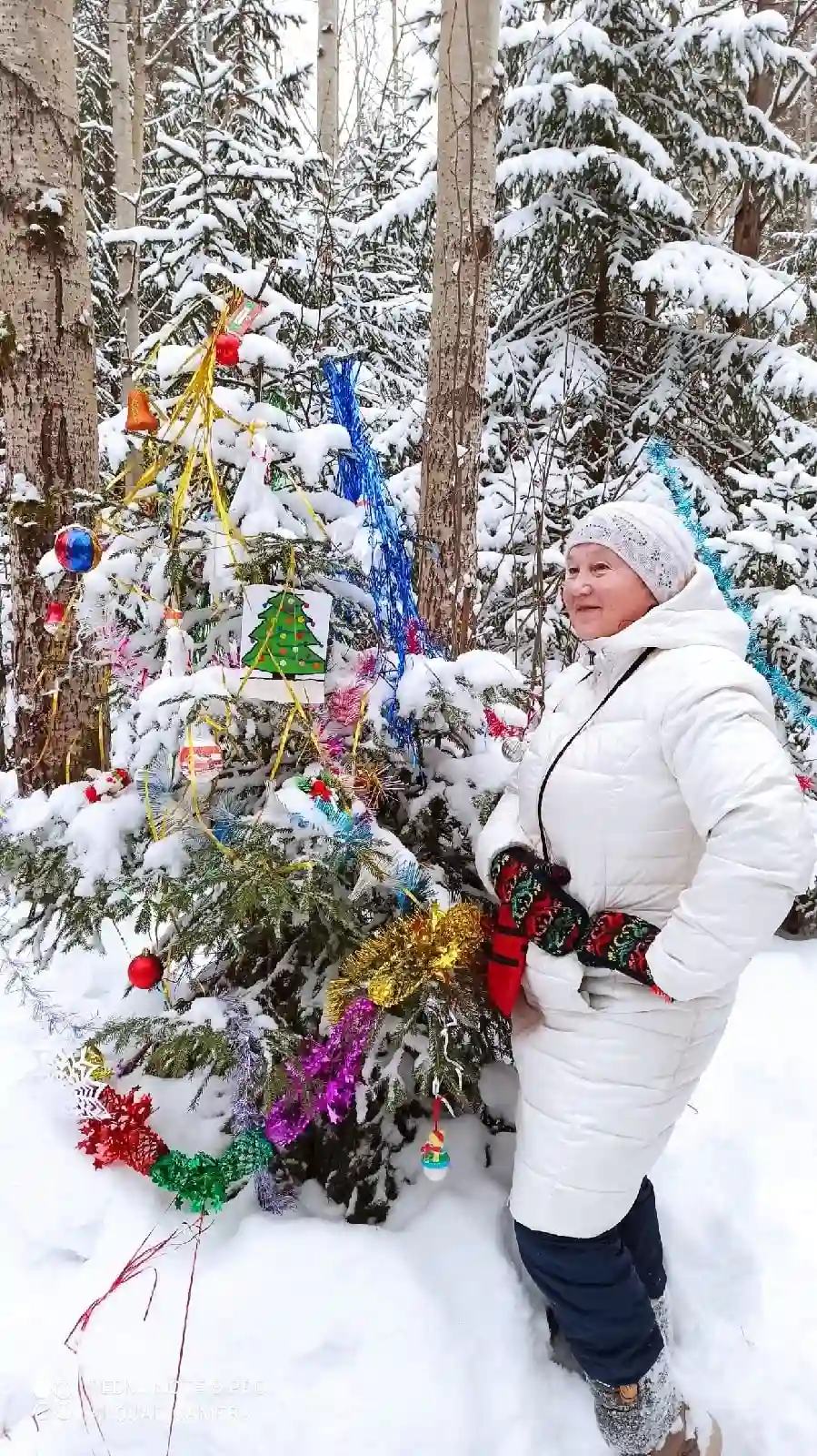 Женщина в белой куртке и шапке стоит рядом с украшенной ёлочкой в лесу